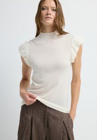 Un haut crème à manches courtes avec des accents volants sur les épaules, présentant un design ajusté et une texture douce, associé à un pantalon marron texturé.