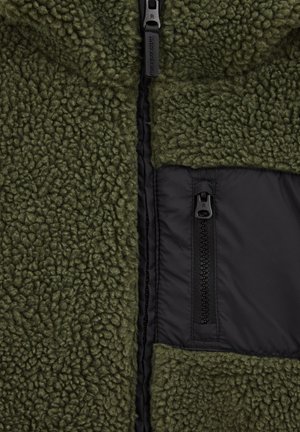 Veste en polaire verte avec un extérieur texturé, une poche zippée noire et une doublure lisse, dotée d'une fermeture éclair à l'avant.
