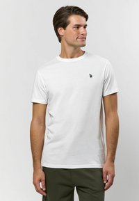 Vit bomulls t-shirt med korta ärmar, rund hals och en liten svart logotyp på vänster bröst. Har ett mjukt, lätt tyg.