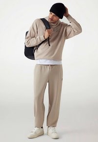 Sudadera beige con el logo "GUESS", combinada con pantalones jogger beige a juego. Lleva un gorro negro y una mochila negra. Zapatillas blancas.