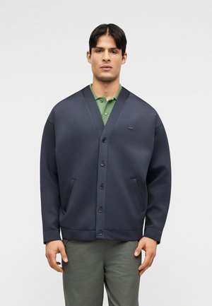 Άνδρας που φοράει σκούρο μπλε ζακέτα με κουμπιά πάνω από πράσινο πόλο πουκάμισο και γκρι παντελόνι, στέκεται μπροστά σε απλό ανοιχτό φόντο.