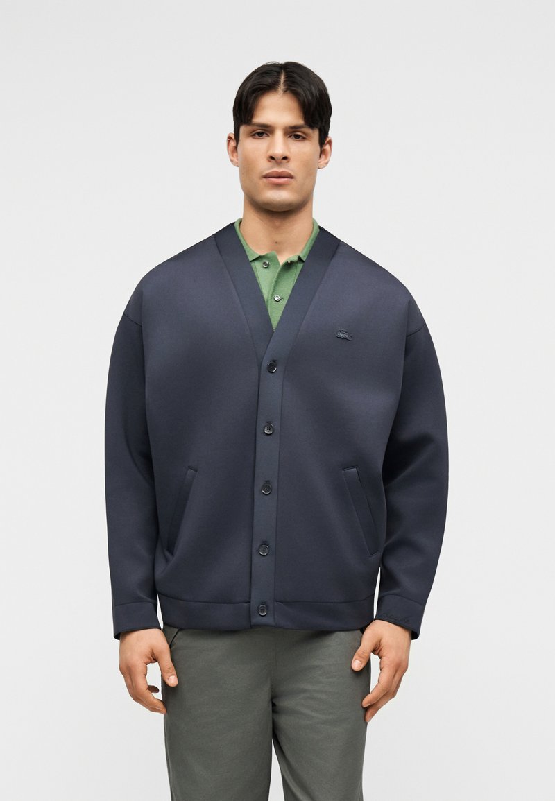 Homme portant un cardigan bleu foncé boutonné sur un polo vert et un pantalon gris, debout devant un fond clair uni.