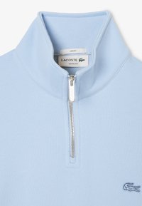 Sudadera de media cremallera en azul claro. Tejido suave con una textura lisa. Presenta una cremallera plateada y un pequeño logo bordado en el pecho.