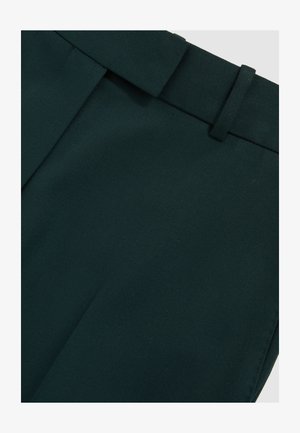 Pantaloni su misura verde scuro con passante per cintura e piega frontale visibile su tessuto liscio.