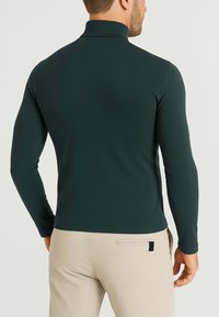 Maglione a maniche lunghe verde scuro con un texture liscia, design aderente e colletto alto. Abbinato a pantaloni chiari.