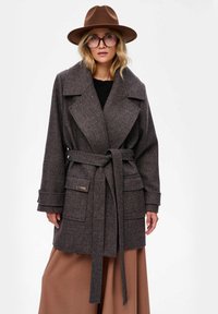 Grijze tekstuur jas met een brede kraag, een ceintuur in de taille en twee voorzakken. Gecombineerd met een bruine hoed en grote brillen.