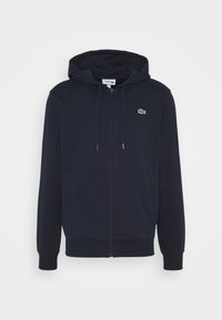 Námořnická modrá mikina na zip vyrobená z bavlněné směsi, s klokaní kapsou, žebrovanými manžetami a malým zeleným logem na levé straně hrudi.