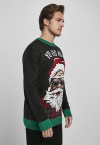 Pulover tricotat negru cu guler și manșete ribelate verzi. Prezintă un grafic cu Moș Crăciun purtând ochelari de soare pe un fundal cu model tartan roșu.