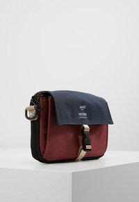 Röd och marinblå axelväska med klaffstängning, svart dragkedja och beige rem, med en slät yta och varumärkeslogotyp. Kompakt rektangulär form.