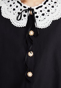 Rochie neagră cu un guler din dantelă albă, având nasturi în formă de floare aurii cu centre din perlă și o panglică la gât.