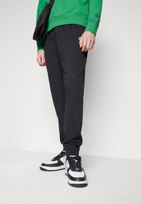 Pantaloni della tuta neri con polsini elastici, abbinati a sneakers bianche e nere. Indossato un felpa verde, completata da una piccola borsa nera.