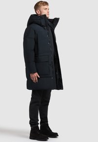 Polstret lang sort frakke med høj krave, store forlommer og trykknapper. Bæres med sorte jeans og snørebåndsstøvler.