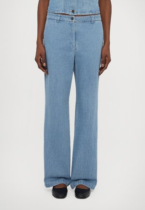 Personne portant un jean taille haute bleu clair à jambes larges et un gilet en jean assorti, associé à des chaussures noires à bout carré, debout droit.