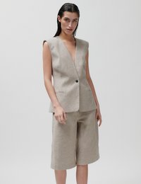 Γυναίκα που φοράει μπεζ αμάνικο σακάκι με μπεζ ρίγες και ένα μαύρο κουμπί, μαζί με αντίστοιχο φαρδύ σορτς μέχρι το γόνατο, στέκεται μπροστά σε απλό φόντο.