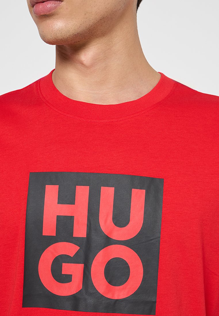 Camiseta de algodón roja con un gran cuadrado negro que lleva el texto "HUGO" en letras rojas en negrita. Cuello redondo y mangas cortas.