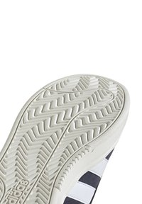 Vit gummisula av sneakers med ett strukturerat mönster och djupa spår; tre horisontella blå och vita ränder över den övre sektionen.