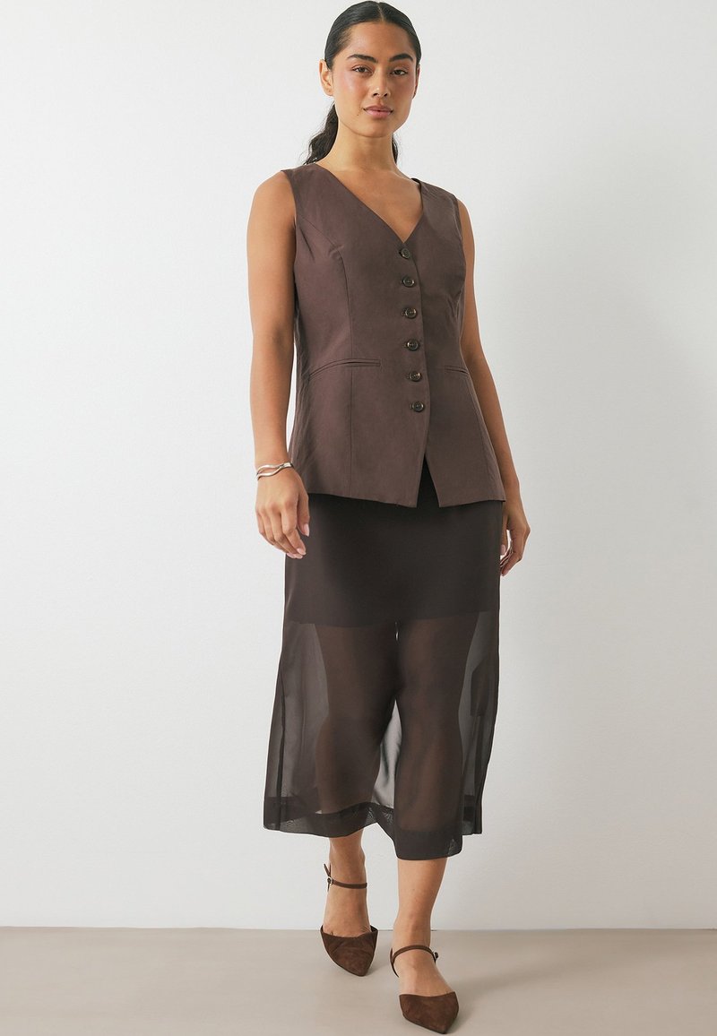 Gilet sans manches marron avec un col en V et une fermeture à boutons, associé à une jupe noire transparente. Des escarpins en suède marron à bout pointu complètent le look.