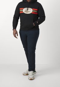 Svart huvtröja med röda och gula randiga accenter, med en logotyp. Kombinerad med mörkblå byxor och vita sneakers med gröna detaljer.