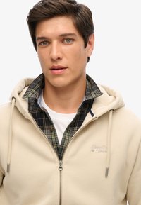 Sudadera beige con cremallera y capucha ajustable, llevada sobre una camisa de cuadros en negro y verde. Presenta un sutil logo en el pecho.