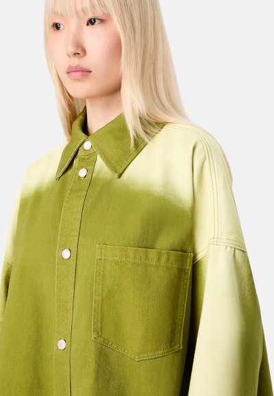 Young woman with long blonde hair wearing oversized green and light yellow gradient button-up shirt with chest pocket.