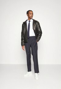 Zwarte leren jas, wit overhemd, donkergrijze broek, marineblauwe stropdas en witte sneakers. Model staat met één hand in de zak.