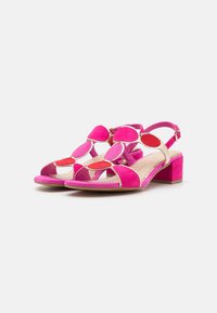 Pink suede sandals featuring a block heel, open toe, and decorative cut-out accents. The design includes circular elements in a contrasting red.