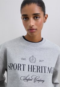 Sudadera gris con cuello redondo. Presenta texto negro "SPORT HERITAGE" y logotipo. Tela de textura suave, ajuste casual y puños acanalados.