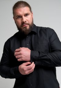 Juoda marškiniai su smailiu apykakle, priekinėmis kišenėmis ir sagčių užsegimu. Ilgos rankovės su sagčių manžetais ir sklandžiu tekstūru.