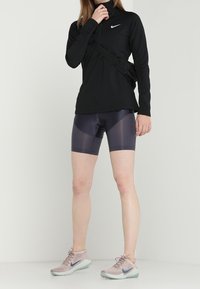 Musta pitkähiha, jossa on vetoketjulla varustettu kaulus ja sivutasku, violetit urheilushortsit ja vaaleanpunaiset juoksukengät, joissa on vaaleansiniset pohjat.