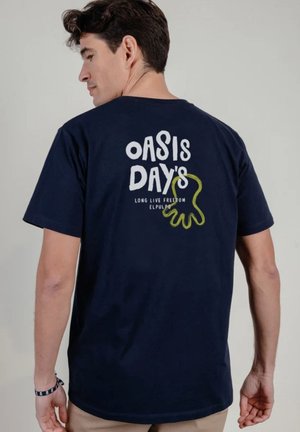Hombre con camiseta azul marino con texto blanco "Oasis Days" y diseño abstracto amarillo en la espalda, mirando hacia su izquierda.