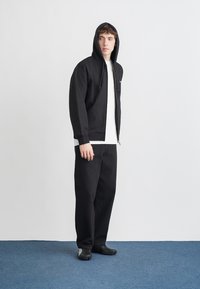 Sudadera negra con cremallera y capucha grande, combinada con una camiseta blanca y pantalones negros de pierna ancha. Calzado negro completa el conjunto.