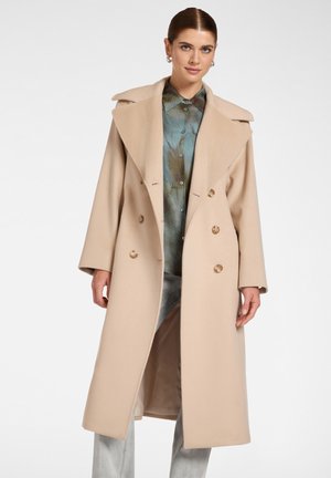 Femme portant un long manteau beige à double boutonnage sur une chemise à motif bleu-vert et un pantalon gris clair, debout sur un fond uni.