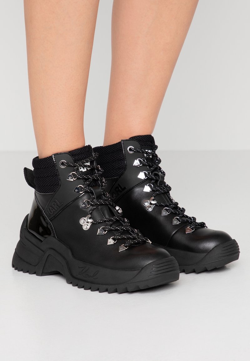 Bottes de randonnée montantes noires avec semelles épaisses, œillets en métal et lacets noirs à motifs portées sur des jambes nues sur un fond gris.