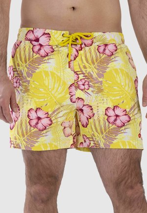 Man wearing yellow swim shorts with pink hibiscus flowers and green tropical leaves, tied with a yellow drawstring at the waist.