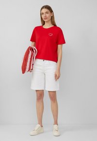 Rotes Baumwoll-T-Shirt mit einem Herz-Motiv, weiße Denim-Shorts und cremefarbene Turnschuhe, hält eine rot-weiß gestreifte Strickdecke.