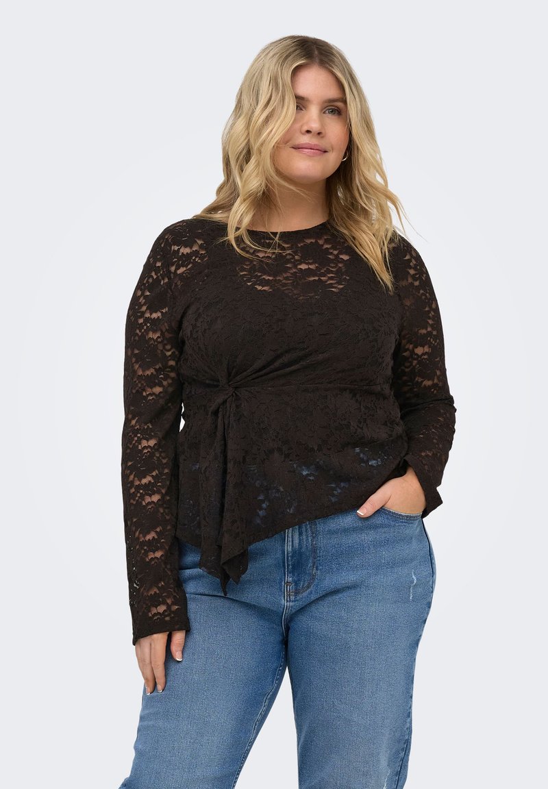 Kvinne med blondt hår iført en svart langermet blonde-topp med en knutedetalj på siden og blå jeans, stående med en hånd i lommen.