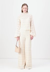 Completo in pizzo floreale a due pezzi in crema, con maniche a campana e pantaloni a gamba larga; abbinato a una piccola borsa beige. Dettaglio in zigzag testurizzato.