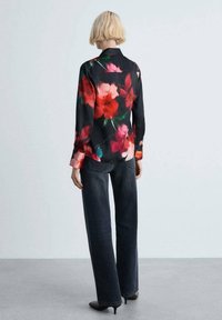 Blouse noire à motifs floraux rouges et verts, manches longues et col, associée à un jean denim foncé à jambes larges et des talons noirs.