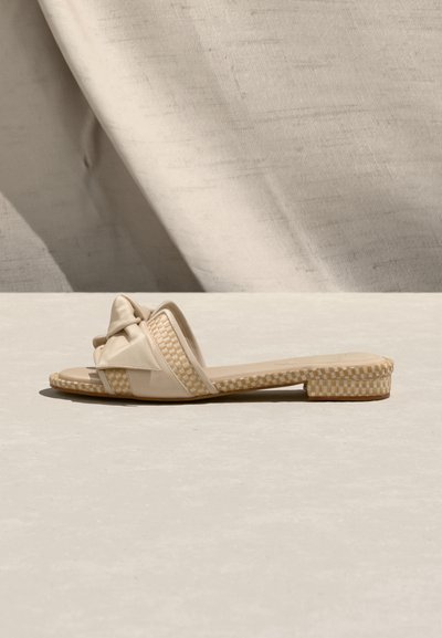 Μπεζ επίπεδη σανδάλι με λουρί που έχει κόμπο και πλεκτές καρό λεπτομέρειες στη σόλα και τη φτέρνα, παρουσιασμένο σε ανοιχτή επιφάνεια με υφασμάτινο φόντο.