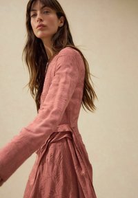 Långärmad, rosafärgad klänning med texturerade bladmönster. Designen har en figursydd överdel och ett plisserat bakstycke.
