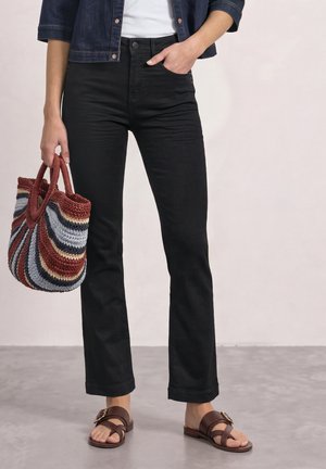 Personne portant un jean noir, une veste en jean, des sandales marron, et tenant un sac fourre-tout tricoté rayé avec une main dans la poche.