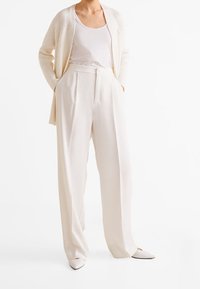 Cardigan créme à texture côtelée, superposé à un débardeur blanc, associé à un pantalon large créme et des chaussures à bout pointu gris clair.