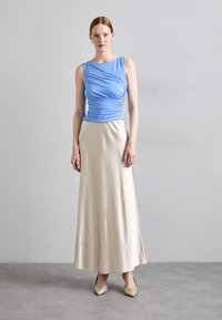 Haut sans manches bleu avec un détail froncé, associé à une jupe beige longue jusqu'au sol. Le modèle porte des chaussures dorées à bout pointu. Fond simple.