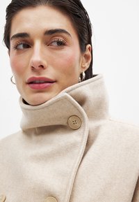 Beige fleece jas met een hoge kraag en twee grote ronde knopen. Gladde textuur en strakke lijnen die een minimalistisch ontwerp benadrukken.