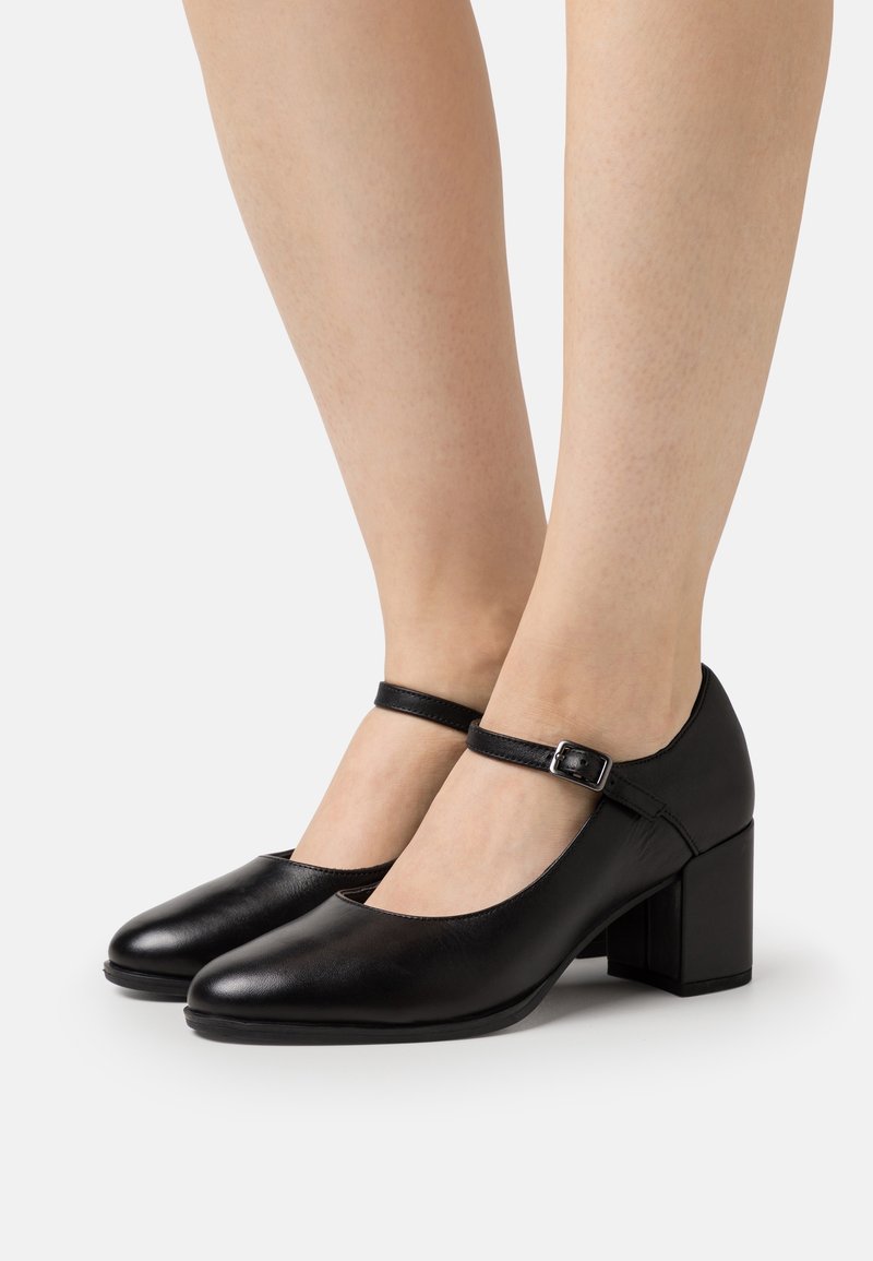 Chaussures Mary Jane en cuir noir avec talons carrés et brides à la cheville, portées pieds nus sur un fond blanc uni.