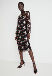 Vestido preto com estampado floral, apresentando flores bordadas em rosa e branco, mangas longas transparentes, gola alta e um silhueta ajustada. Usado com saltos pretos.