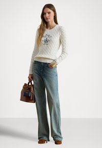 Ženska nosi belo pulover z vzorcem kabel, modre ohlapne jeans hlače, rjave pete in nosi rjavo torbico z majhno mornarsko modro torbico in okrasnimi cofki.