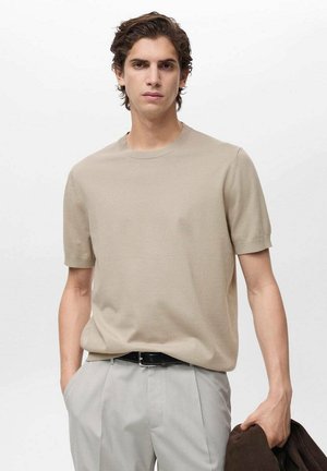 T-shirt beige a maniche corte in maglia con scollatura rotonda, mostrato insieme a pantaloni grigi e una giacca marrone scuro, con texture liscia.