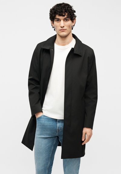 Νέος άνδρας με σγουρά μαλλιά φοράει μαύρο μακρύ παλτό πάνω από λευκή μπλούζα και ανοιχτόχρωμα μπλε τζιν, στέκεται μπροστά σε απλό φόντο.