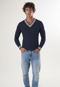 Pullover in maglia a treccia di colore navy con scollatura a V e accenti a righe bianche su colletto e polsini, abbinato a jeans azzurri chiari. Design semplice e testurizzato.